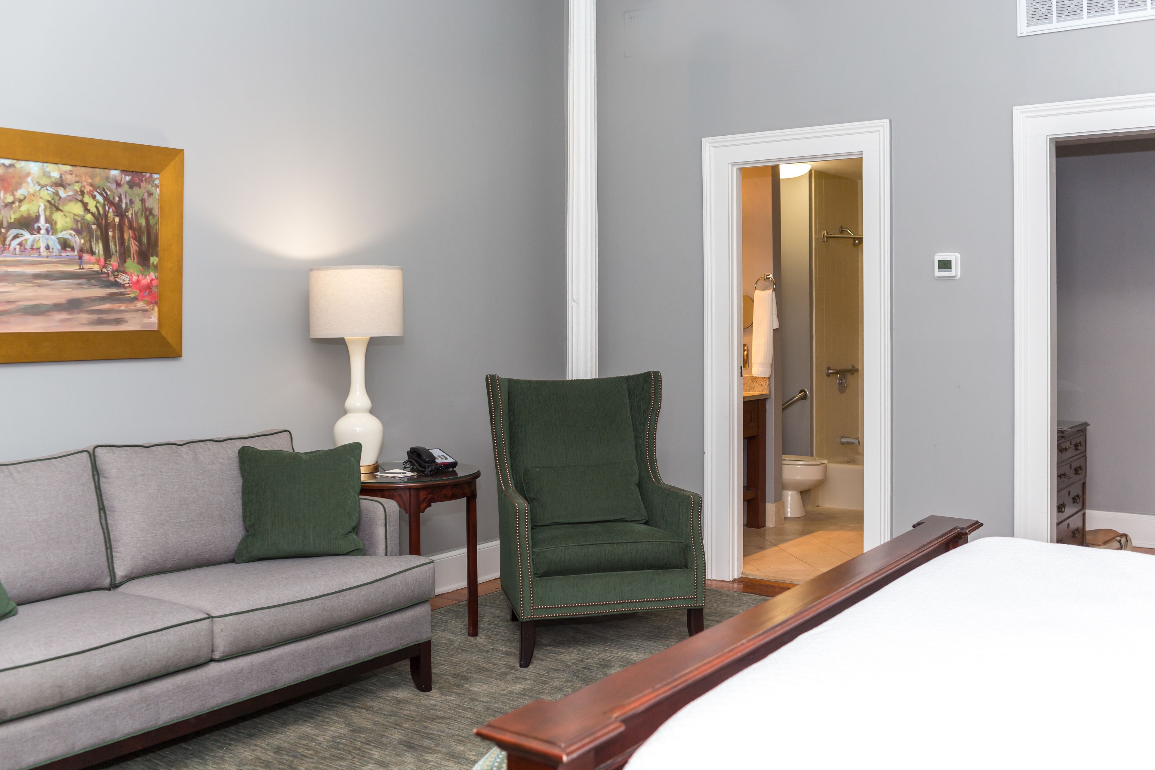 A hotel room with a bed, a green armchair, a grey sofa, a lamp, and a painting on the wall.