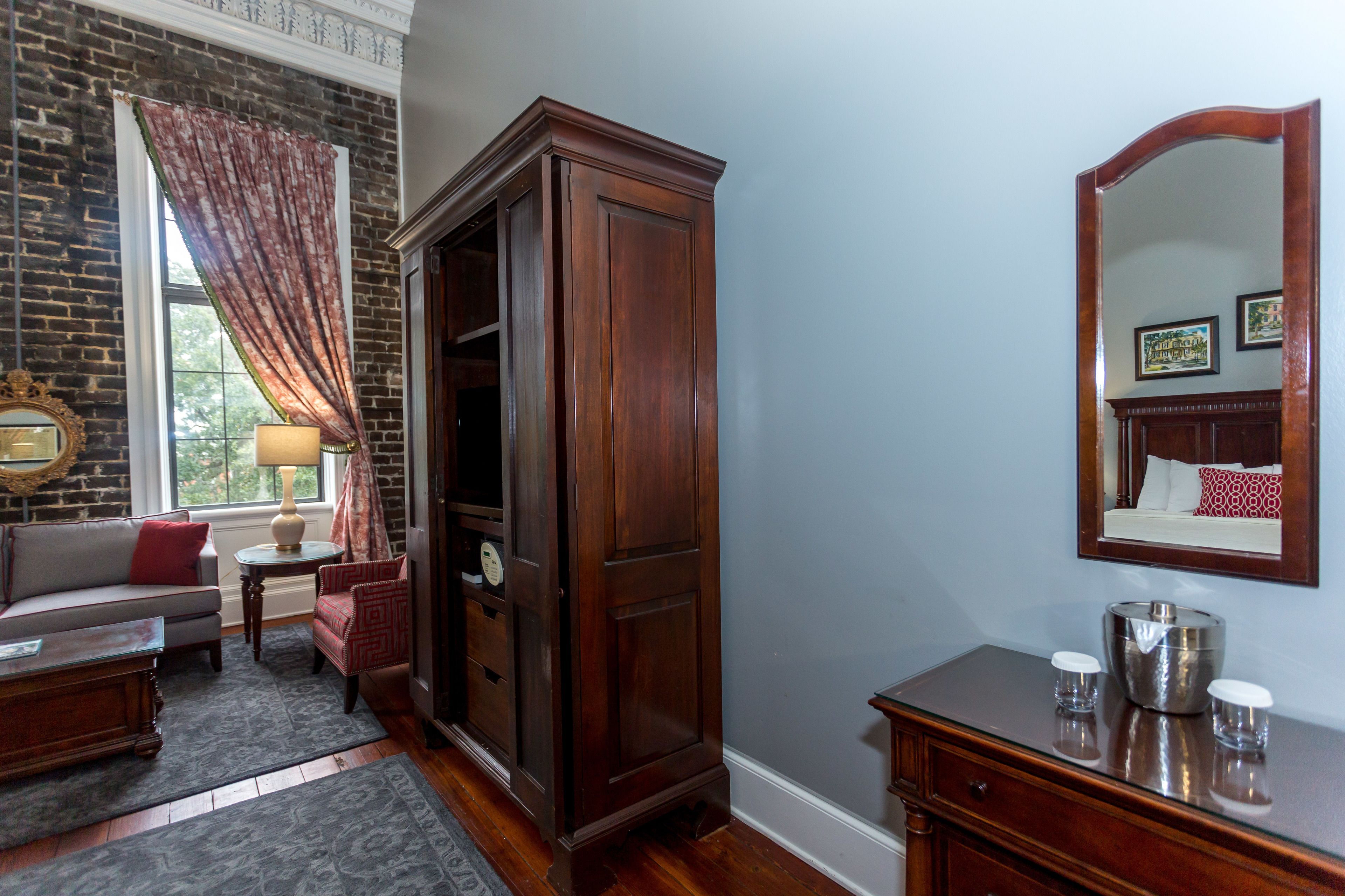 A room with a brick wall, a wooden wardrobe, a mirror, a sofa, a small table, and a lamp.