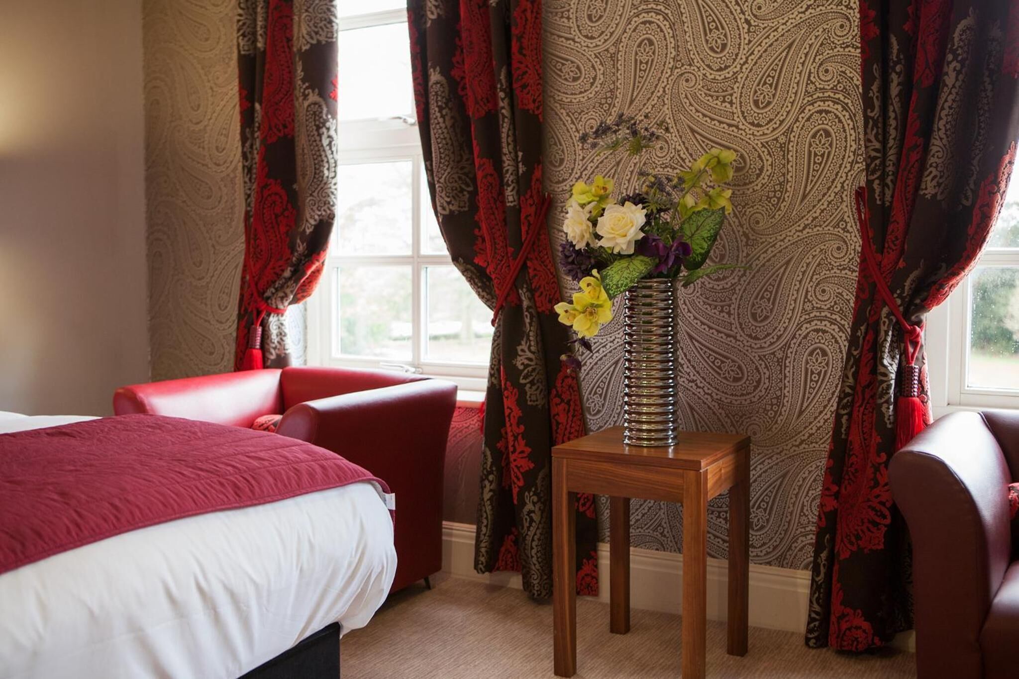 Una habitación de hotel con una cama, una silla roja, una mesita de madera con un jarrón de flores y cortinas estampadas.
