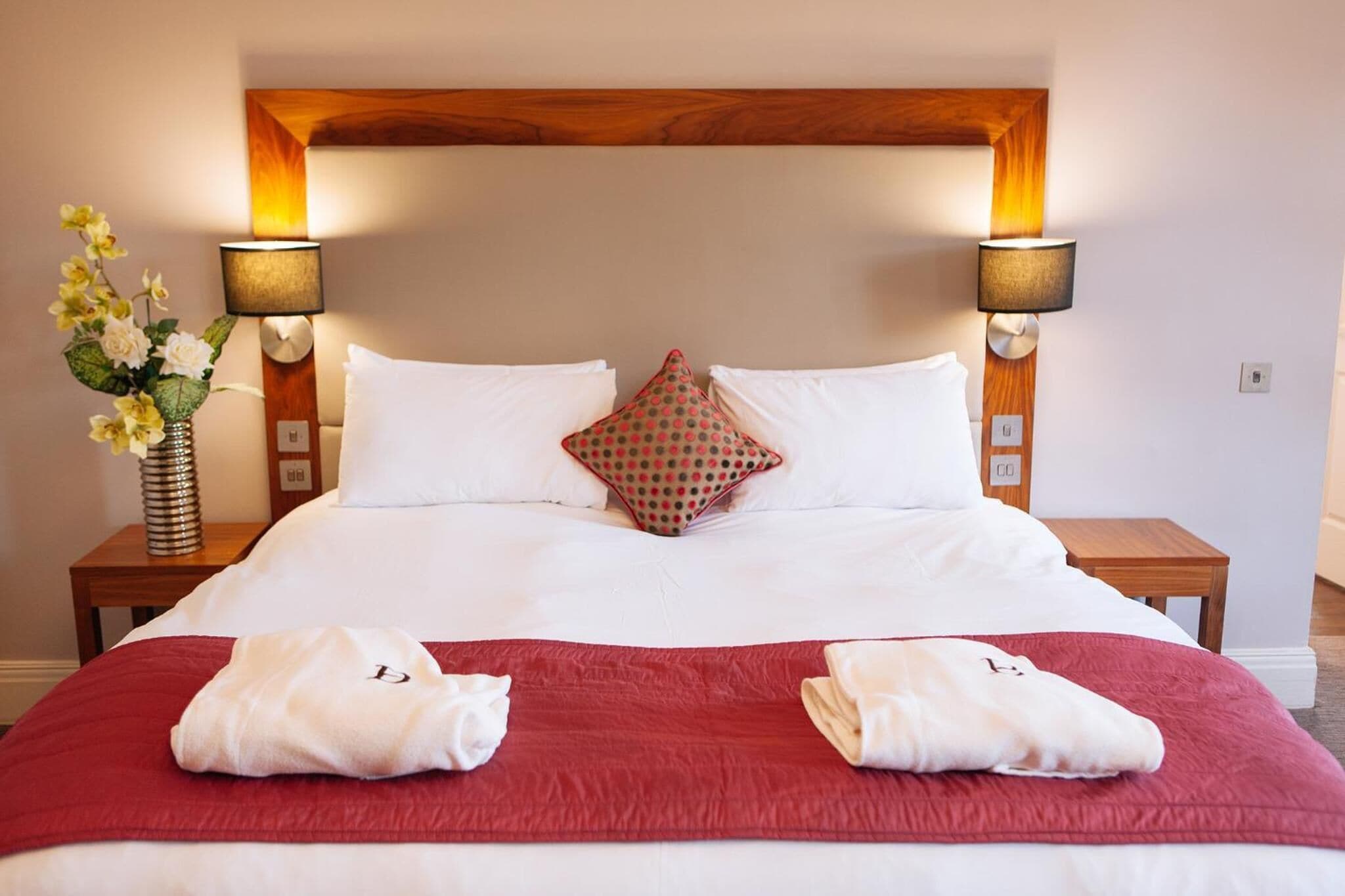 A hotel room with a large bed, two bedside lamps, a wooden headboard, and a red throw blanket.
