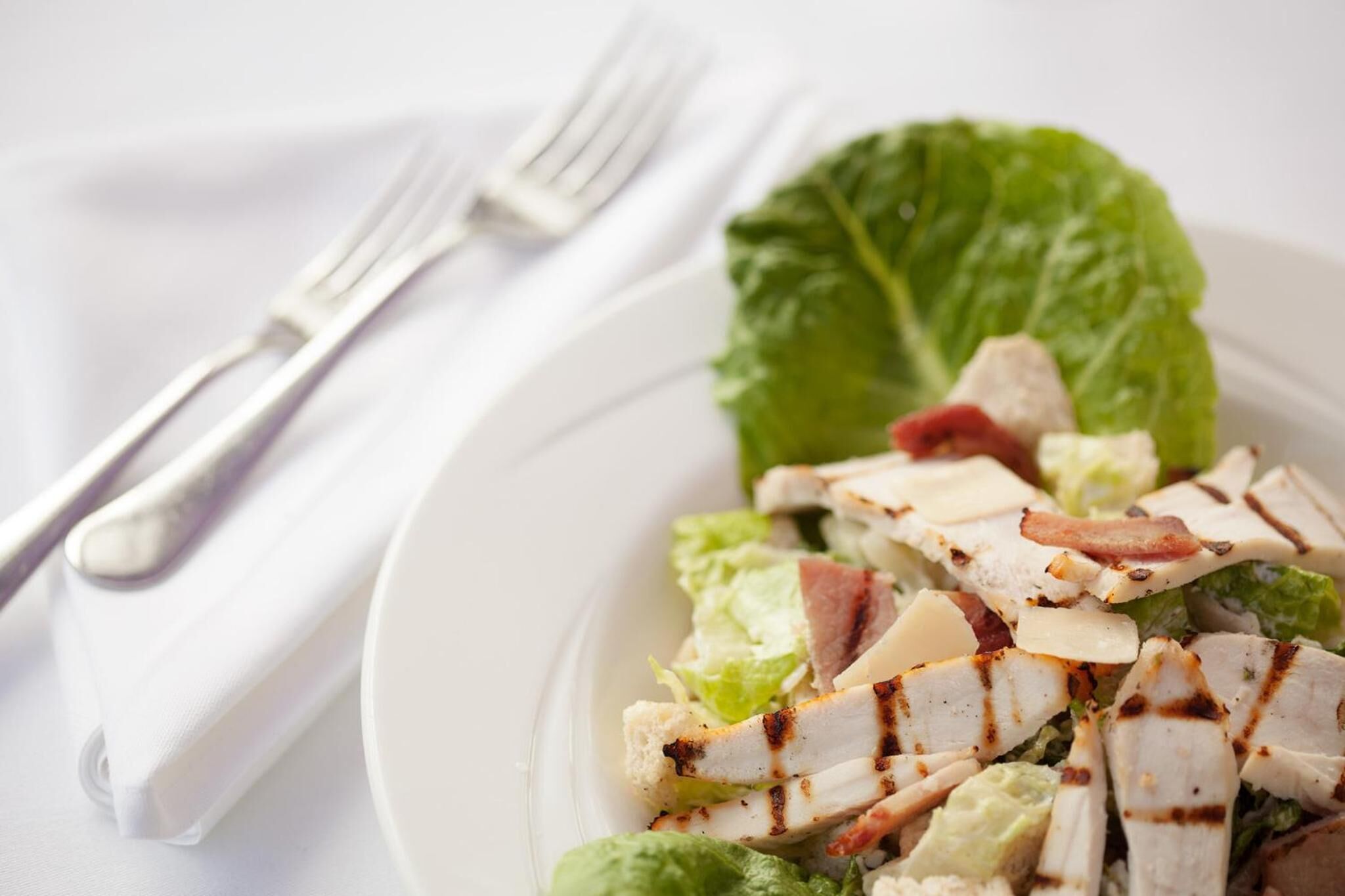 Prato de salada de frango grelhado com alface e bacon, acompanhado de um garfo e guardanapo sobre uma toalha de mesa branca.
