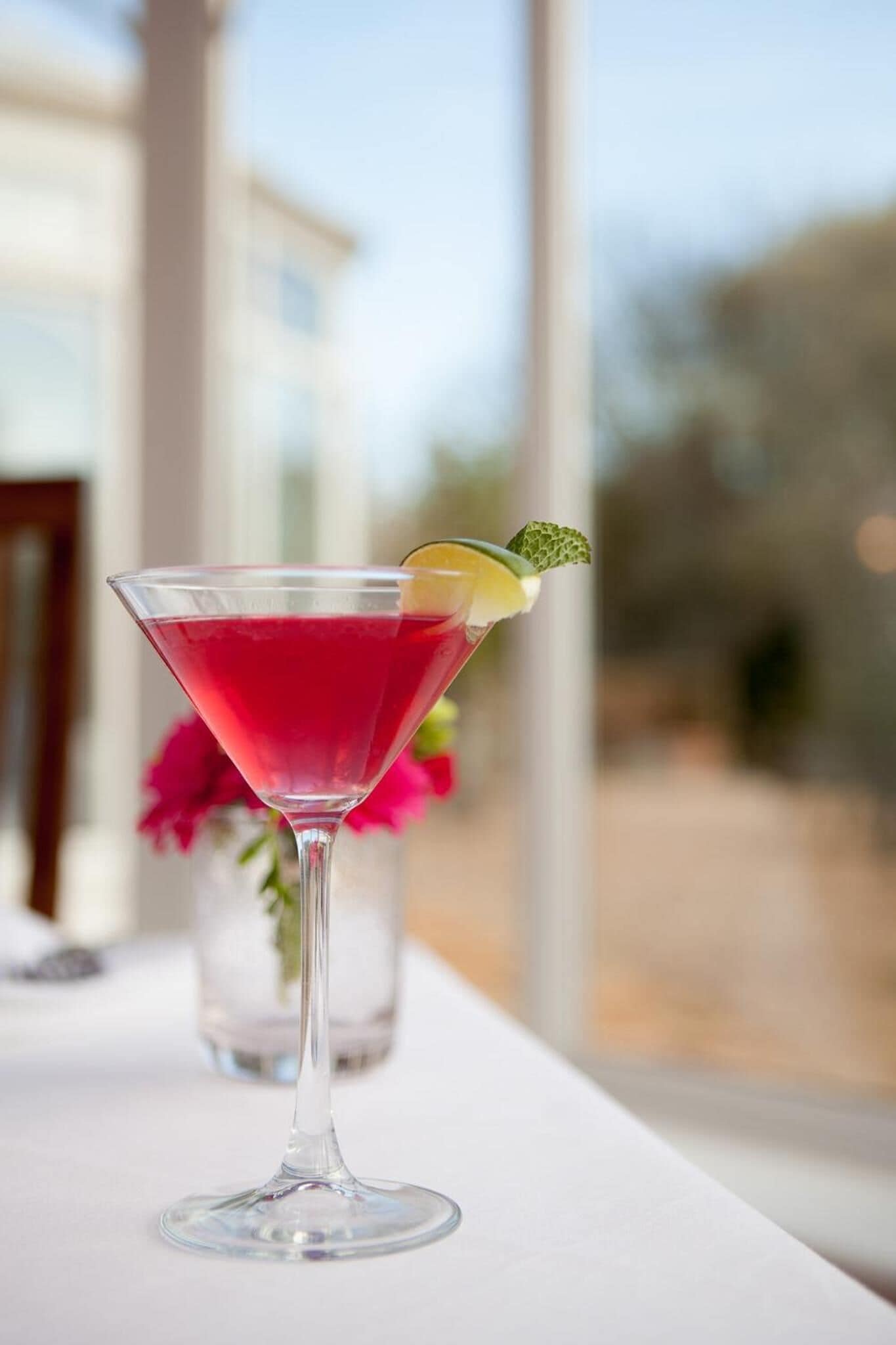 A martini glass with a red drink, garnished with a lime wedge and mint, on a white table with a blurred background of a building and greenery.