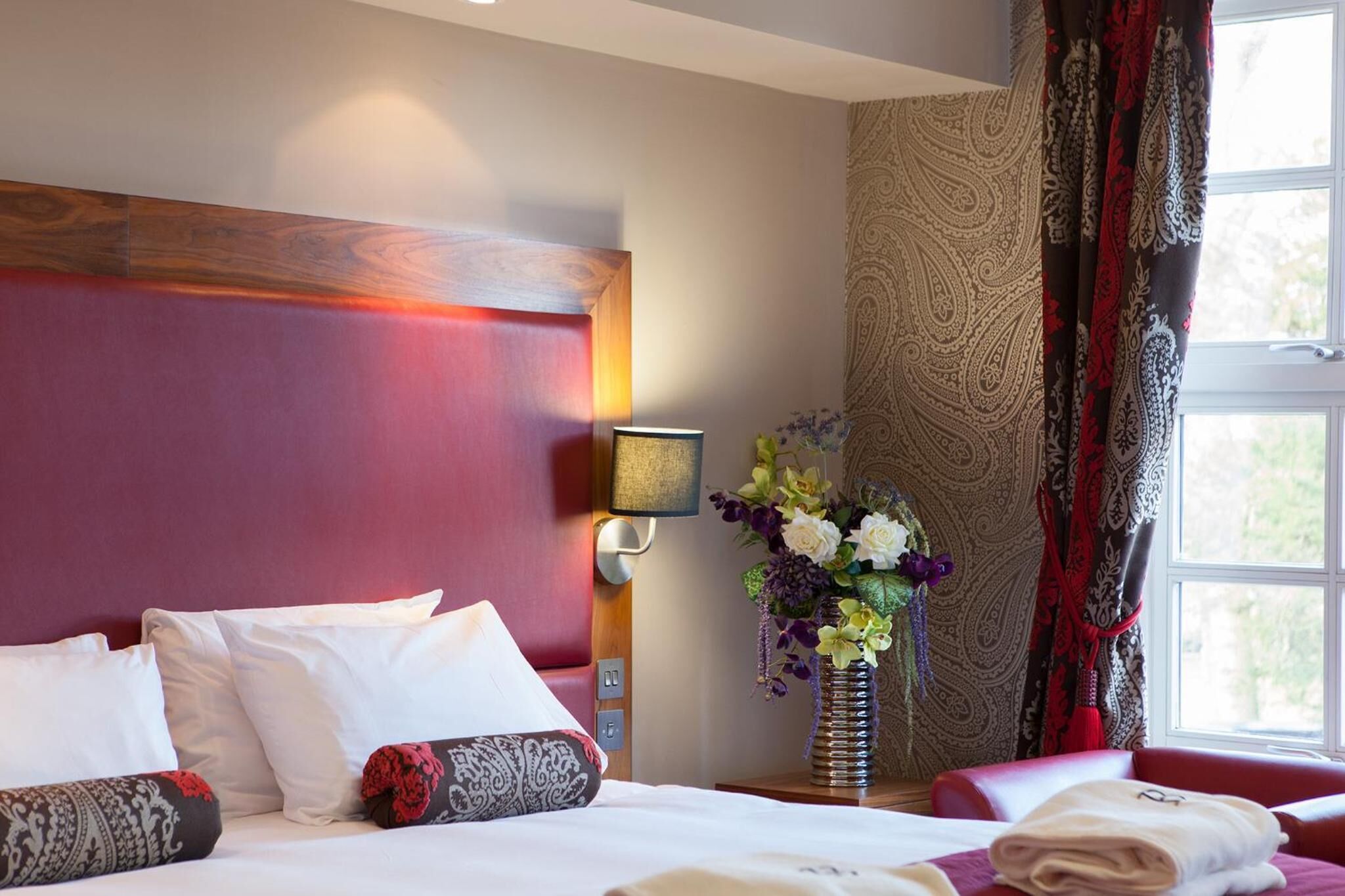 Habitación de hotel con una cama grande, una mesita de noche con un jarrón de flores, una lámpara de pared y una ventana con cortinas.