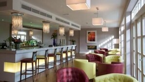 Un área de bar moderna con una barra larga y varios taburetes. Varias lámparas colgantes y un ventanal grande que ofrece luz natural.