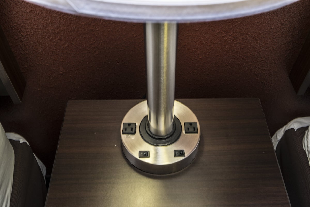 A metallic table lamp with a white shade, a circular base with two electrical outlets, and a metallic stand.