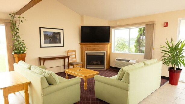 A living room with two green sofas, a wooden coffee table, a fireplace, a TV, and a large window with curtains. - Royal Inn & Suites (Milaca)