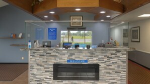 A reception area with a 'WELCOME' sign, a reception desk, a computer, and a window with a view of the outdoors. - Royal Inn & Suites (Milaca)