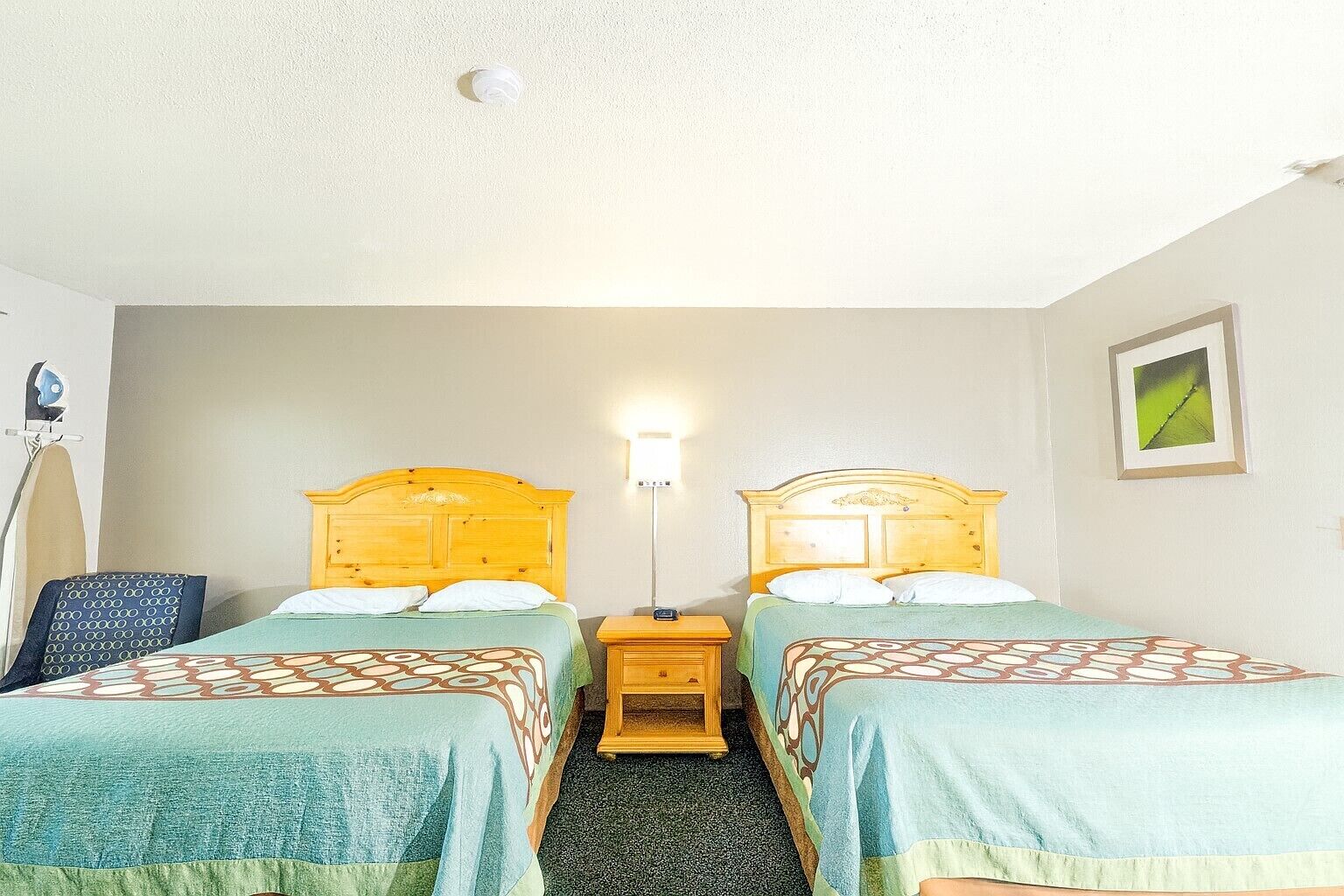 A hotel room with two beds, a nightstand, a lamp, an ironing board, and a framed picture on the wall.