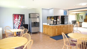 A modern kitchen area with a refrigerator, coffee machine, and a Dr Pepper dispenser. There are wooden cabinets and a dining area with tables and chairs. - Royal Inn & Suites (Milaca)