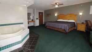 A hotel room with a large bathtub, a bed with a wooden headboard, a bedside table, and a chair. - Royal Inn & Suites (Milaca)