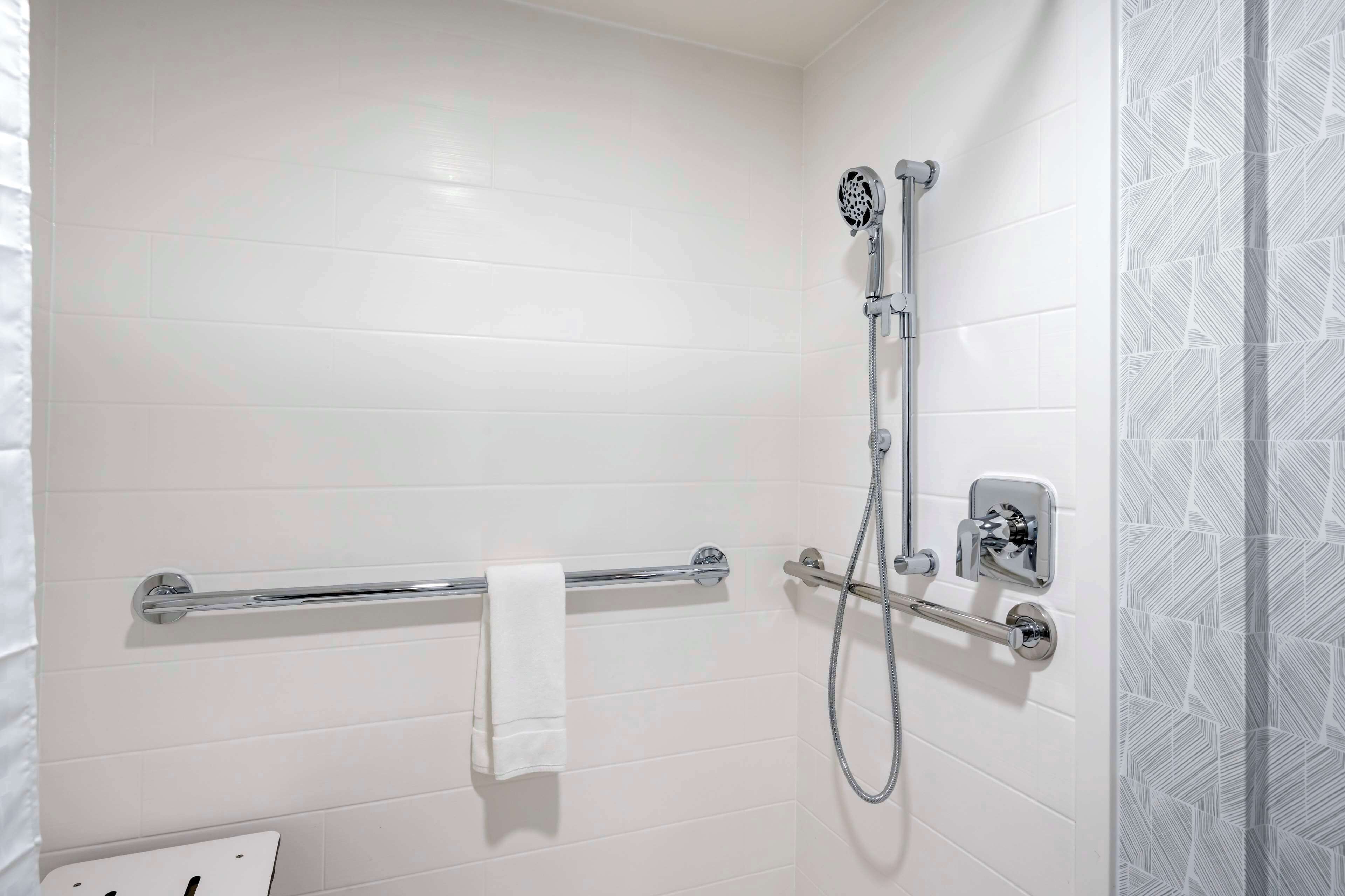 Un baño con paredes de azulejos blancos, una ducha con cabezal de ducha manual, un toallero y un dispensador de papel higiénico.