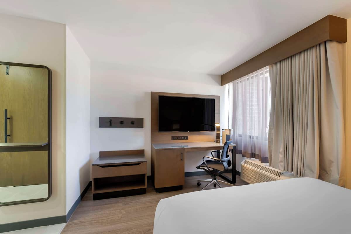 Una habitación de hotel con cama, escritorio con televisor, silla y ventana con cortinas.