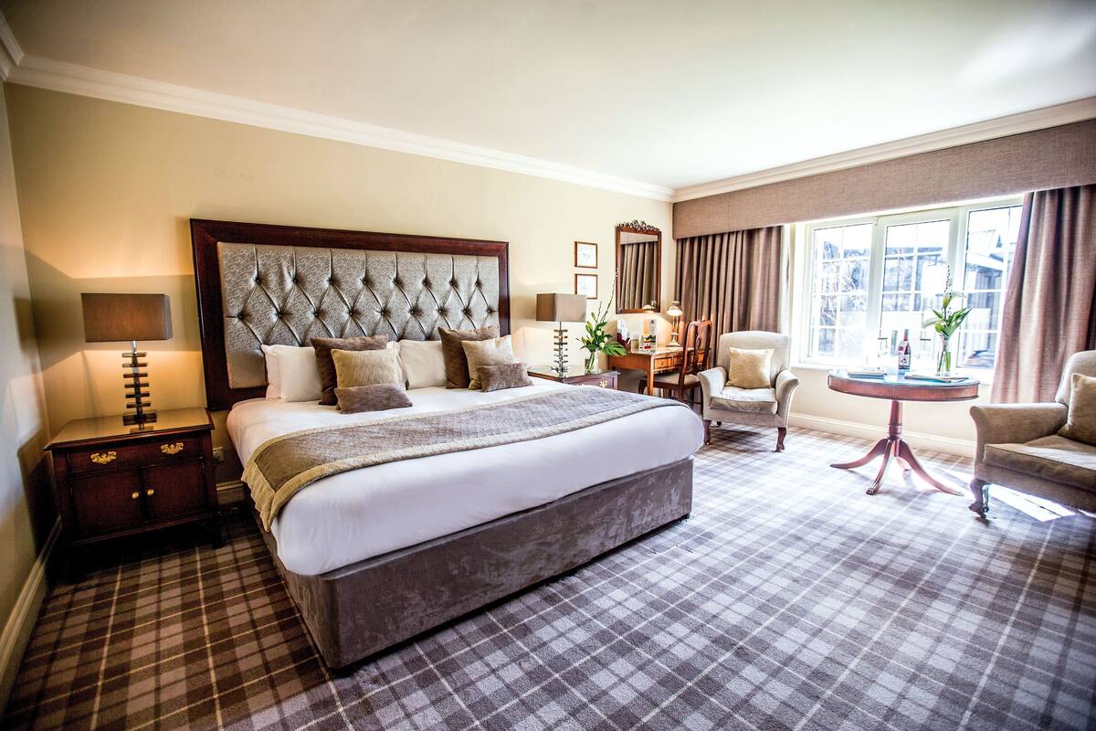 A hotel room with a large bed, a desk, two chairs, and a window with curtains.