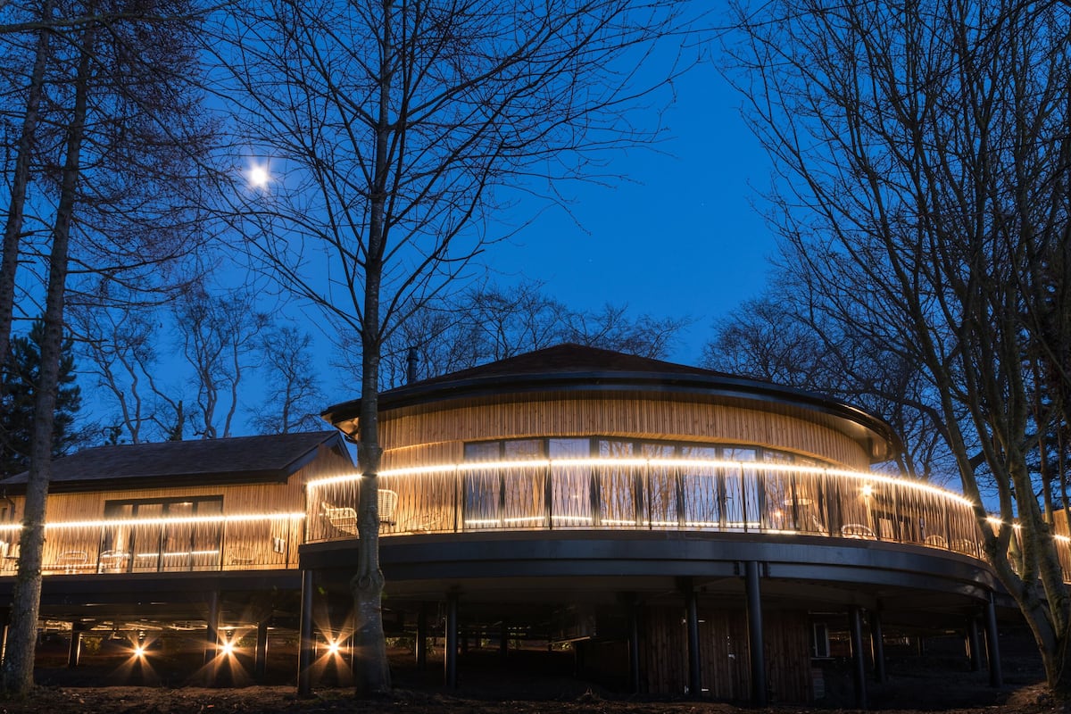 A modern, circular building with a balcony and illuminated interior lights, surrounded by trees.