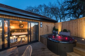 A modern outdoor hot tub area with a wooden deck, a glass-enclosed bedroom with a bed and seating area, and a view of the surrounding trees.