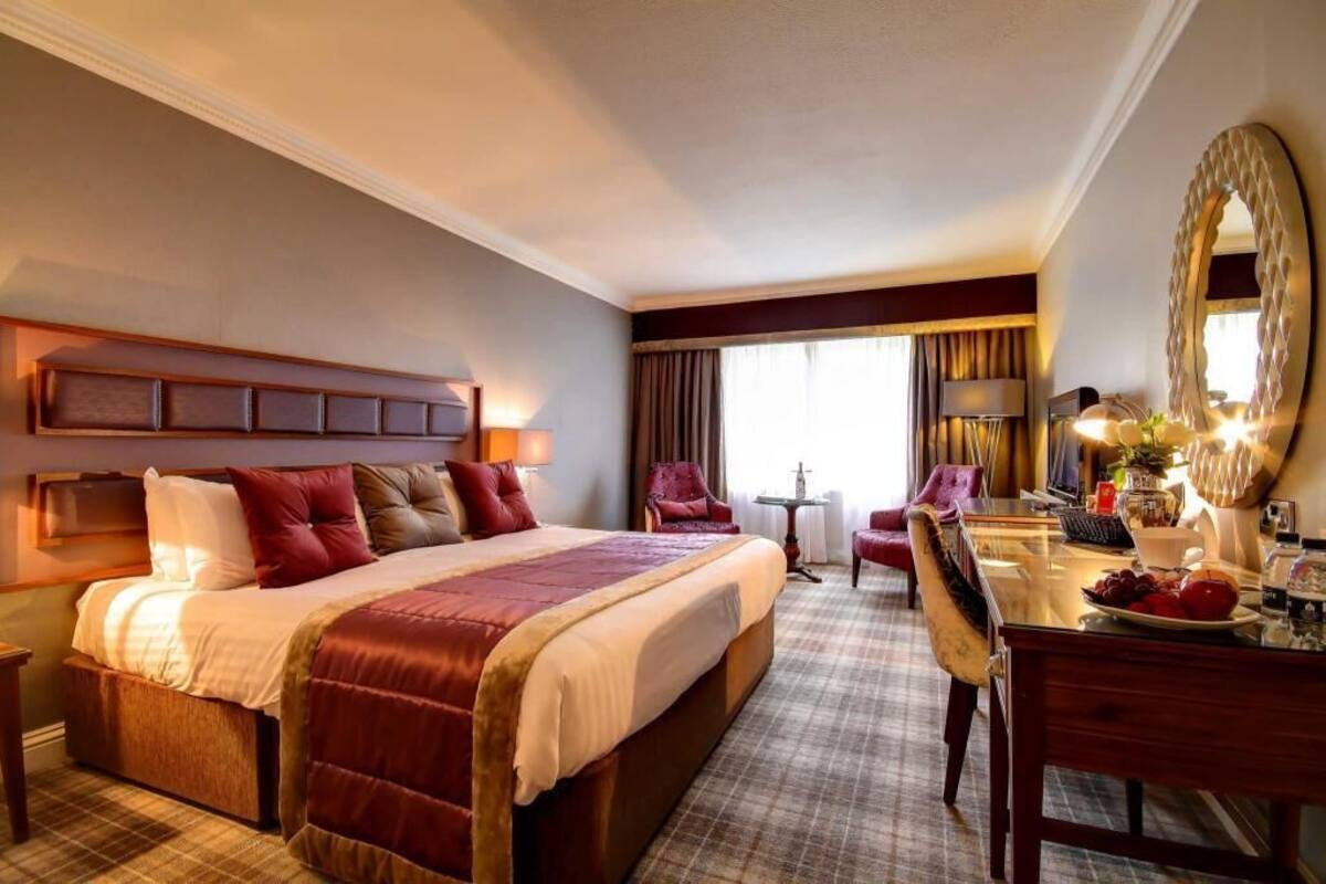 A hotel room with a large bed, a desk with a chair, a mirror, and a window with curtains.