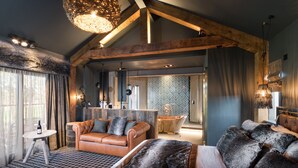 A modern interior with a high ceiling, exposed wooden beams, a cozy seating area with a leather sofa, a large window with sheer curtains, and a visible bathroom with a patterned wall. - Ramside Hall Hotel, Golf and Spa (Durham)