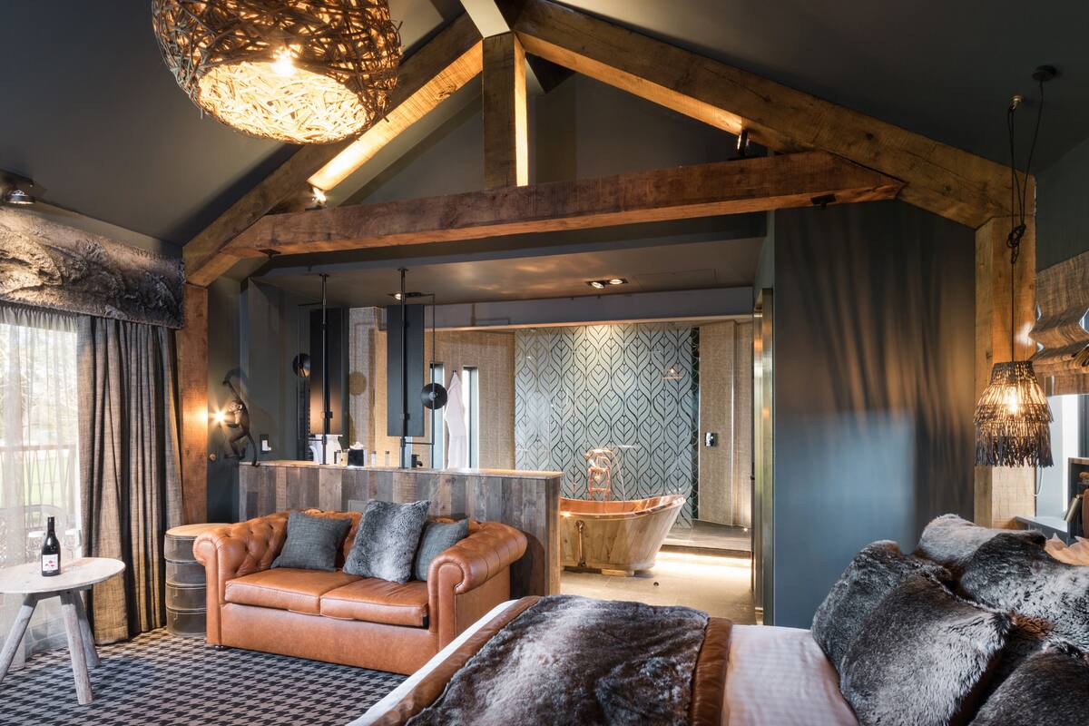 A modern interior with a high ceiling, exposed wooden beams, a cozy seating area with a leather sofa, a large window with sheer curtains, and a visible bathroom with a patterned wall.