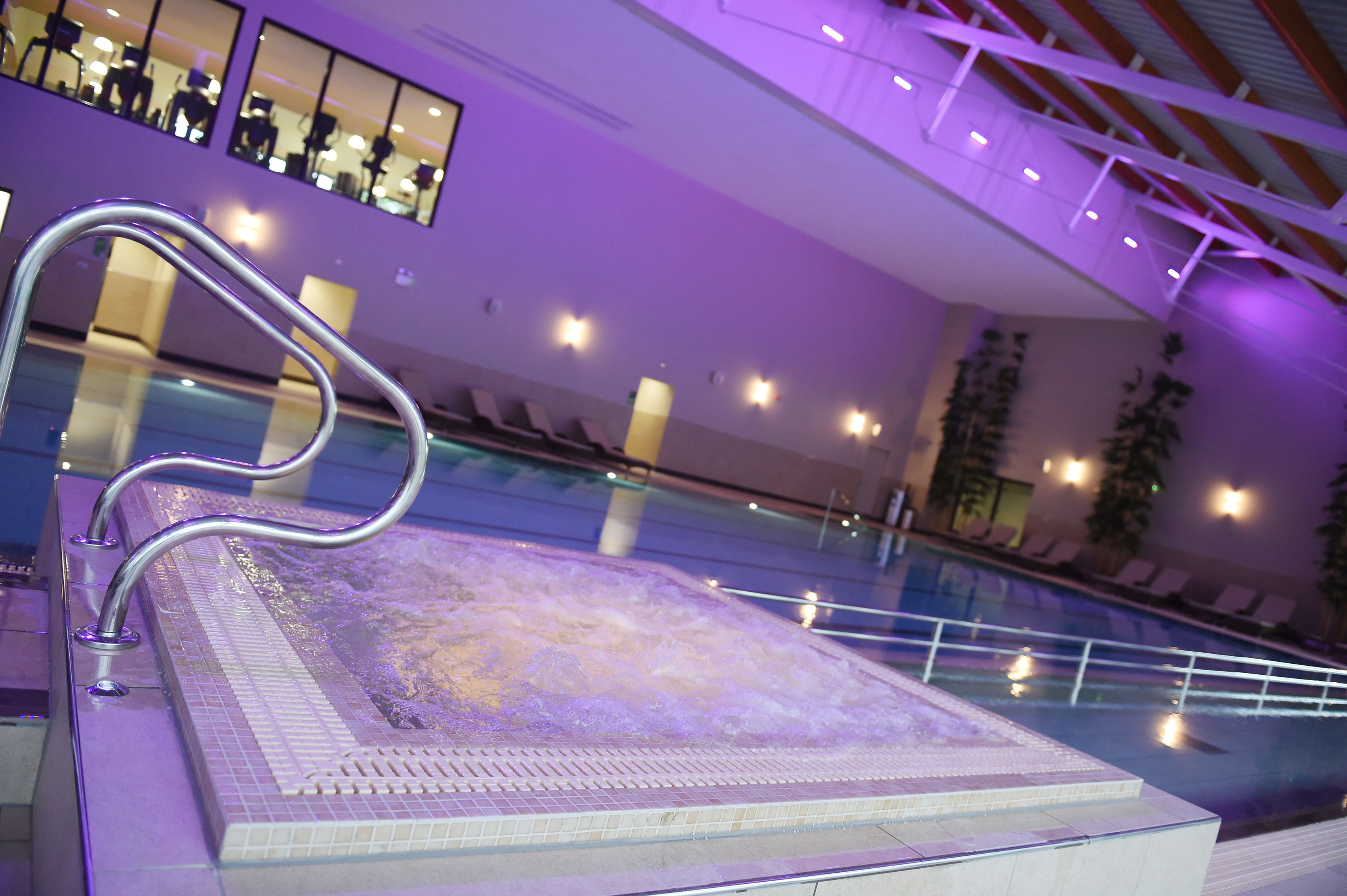 An indoor swimming pool with a tiled deck, a metal handrail, and a row of lounge chairs along the poolside.