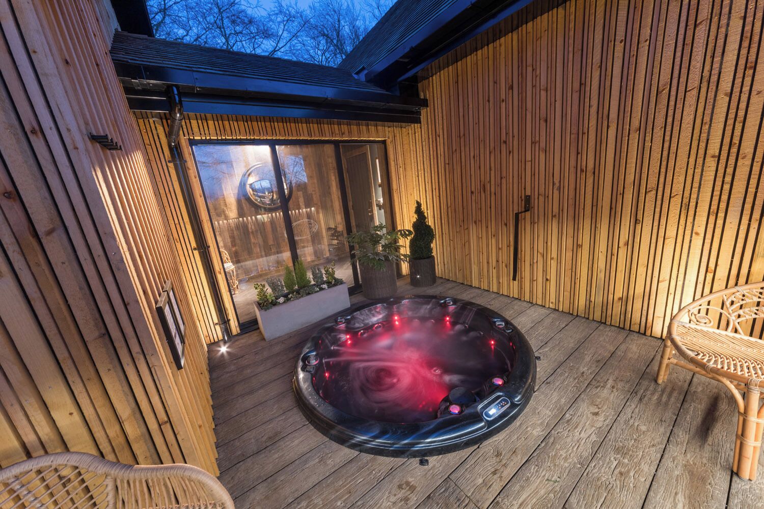 Una terraza de madera con jacuzzi, sillas de mimbre y un pequeño jardín.