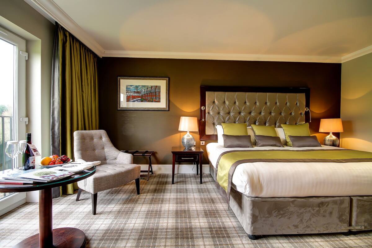 A hotel room with a large bed, a chair, a table with a fruit bowl, and two lamps.