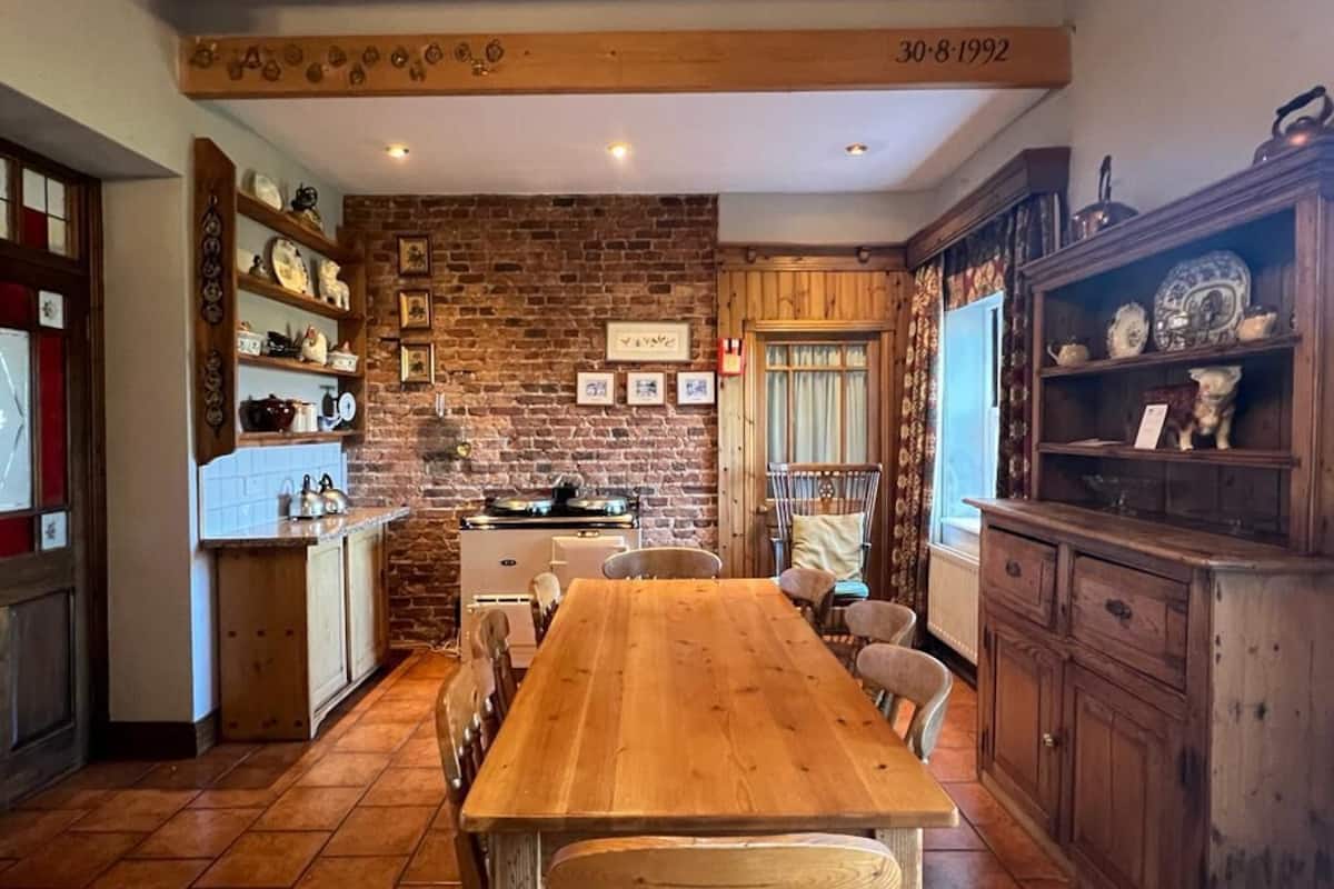 A rustic kitchen with a wooden table, brick wall, and shelves with kitchenware.