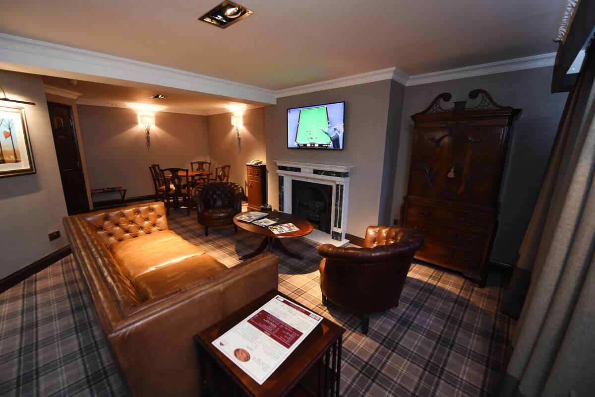 A hotel room with a plaid carpet, leather sofa, armchair, fireplace, television, and a wooden cabinet.