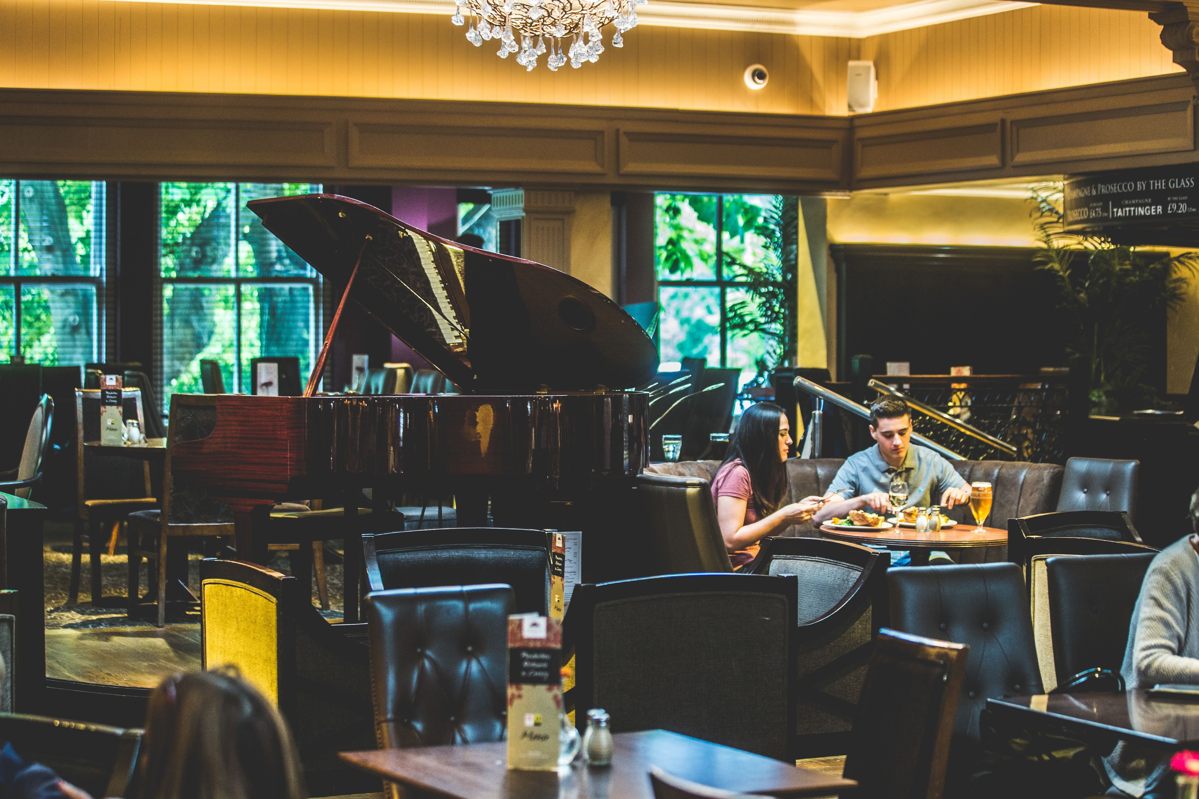 A restaurant with a grand piano, leather seating, and large windows with a view of greenery.
