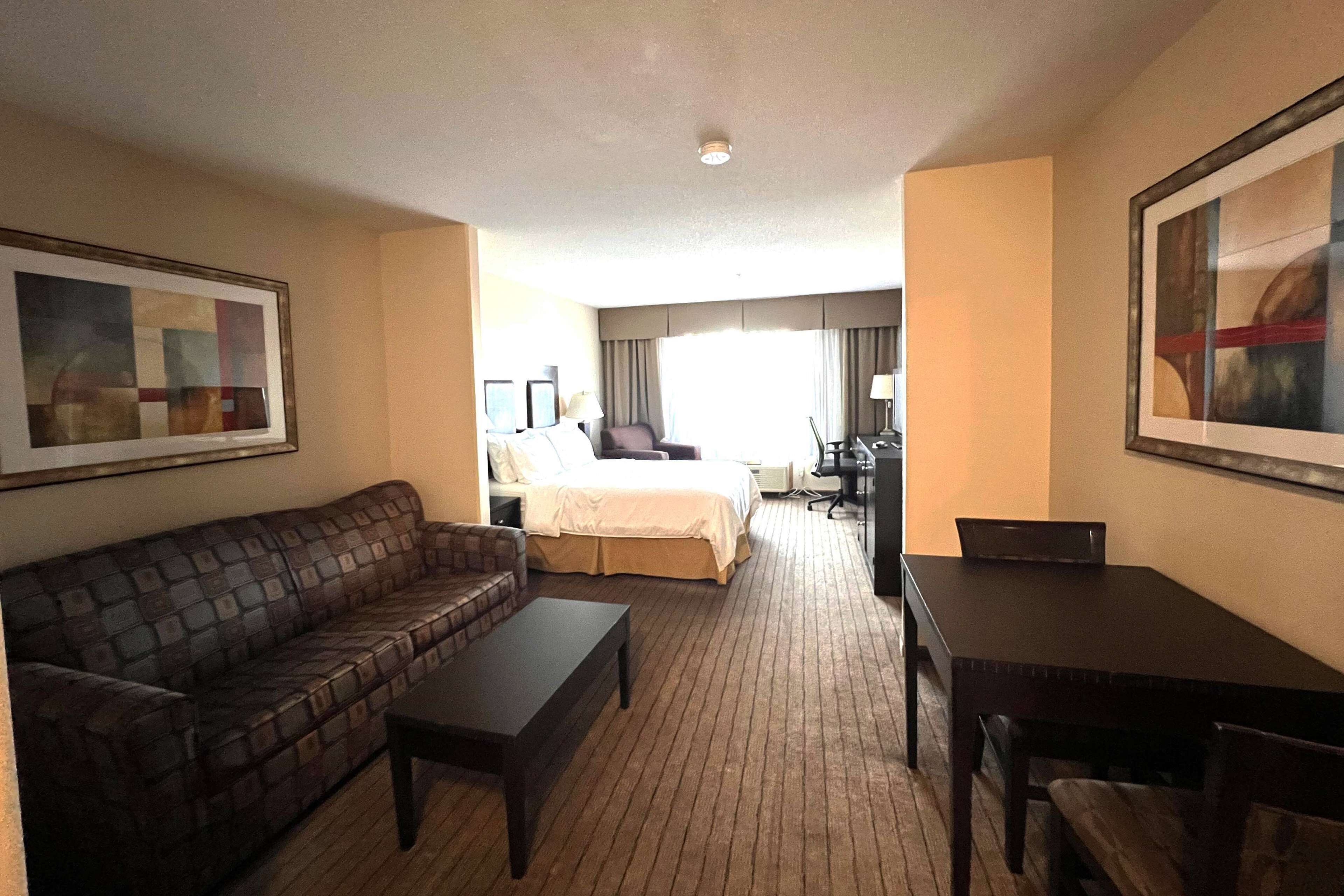 A hotel room with a brown leather sofa, a small table, a bed with a white comforter, a desk with a chair, and a large framed picture on the wall.