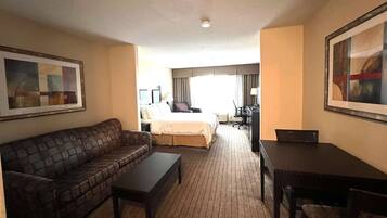 A hotel room with a brown leather sofa, a small table, a bed with a white comforter, a desk with a chair, and a large framed picture on the wall.