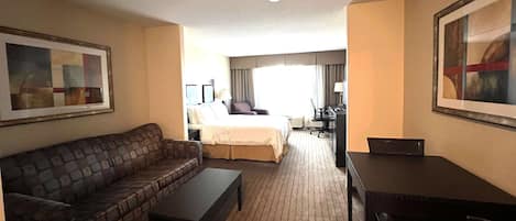 A hotel room with a brown leather sofa, a small table, a bed with a white comforter, a desk with a chair, and a large framed picture on the wall.
