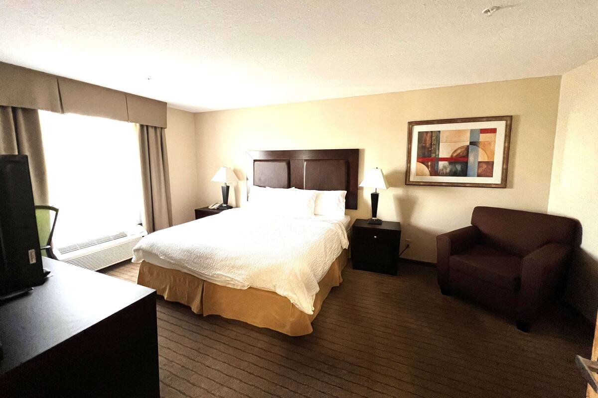 A hotel room with a large bed, two bedside tables with lamps, a chair, a desk with a computer, and a painting on the wall.