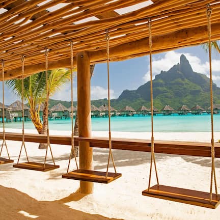 A beach with wooden swings, palm trees, and a view of a mountain and turquoise waters.