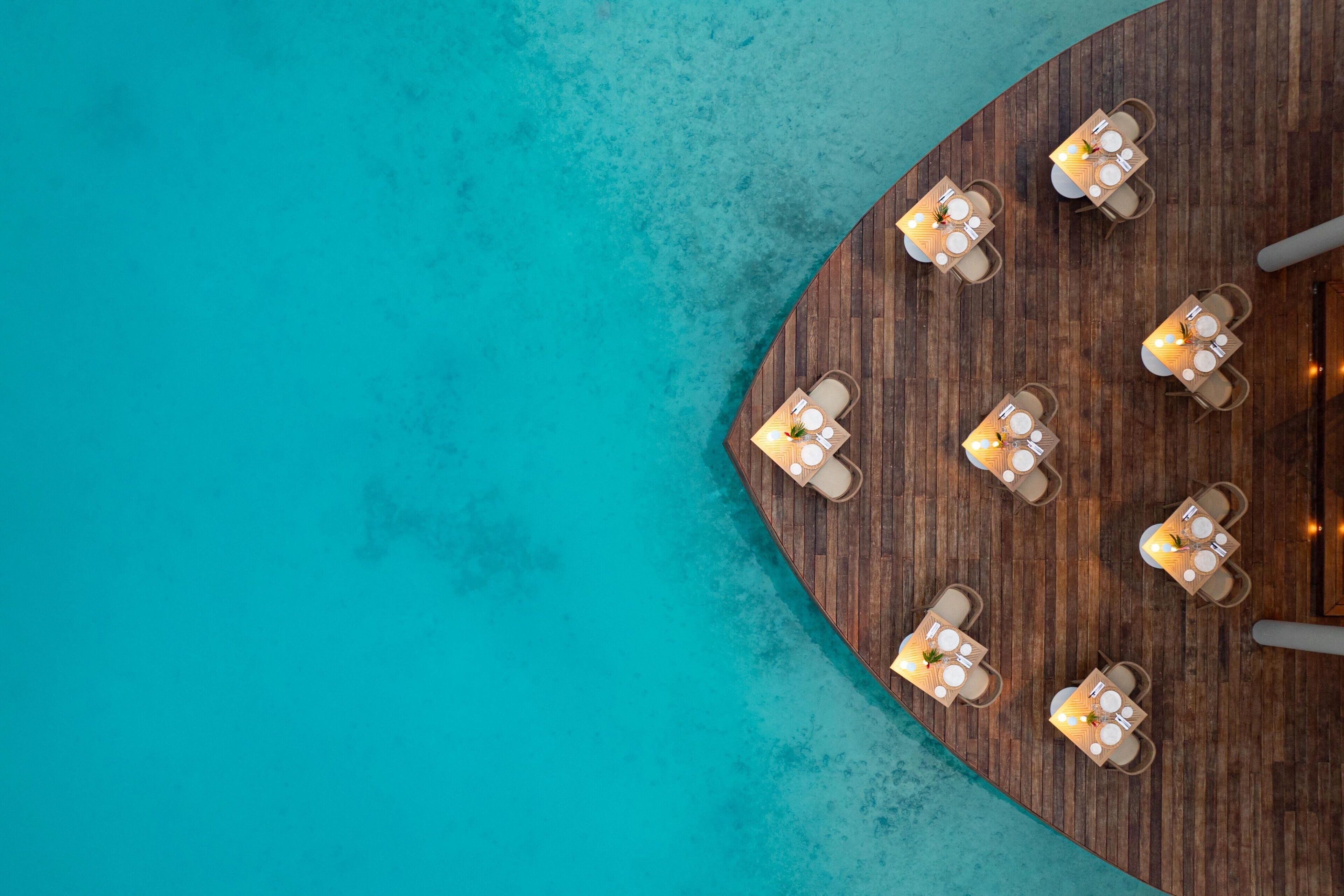 A top-down view of a wooden deck with tables and chairs, overlooking a turquoise sea.