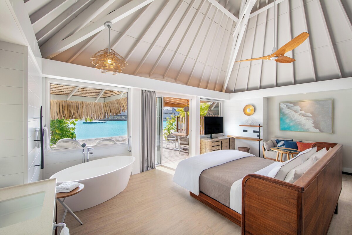 A modern hotel room with a large bed, a bathtub, a dining area, and a view of the pool.