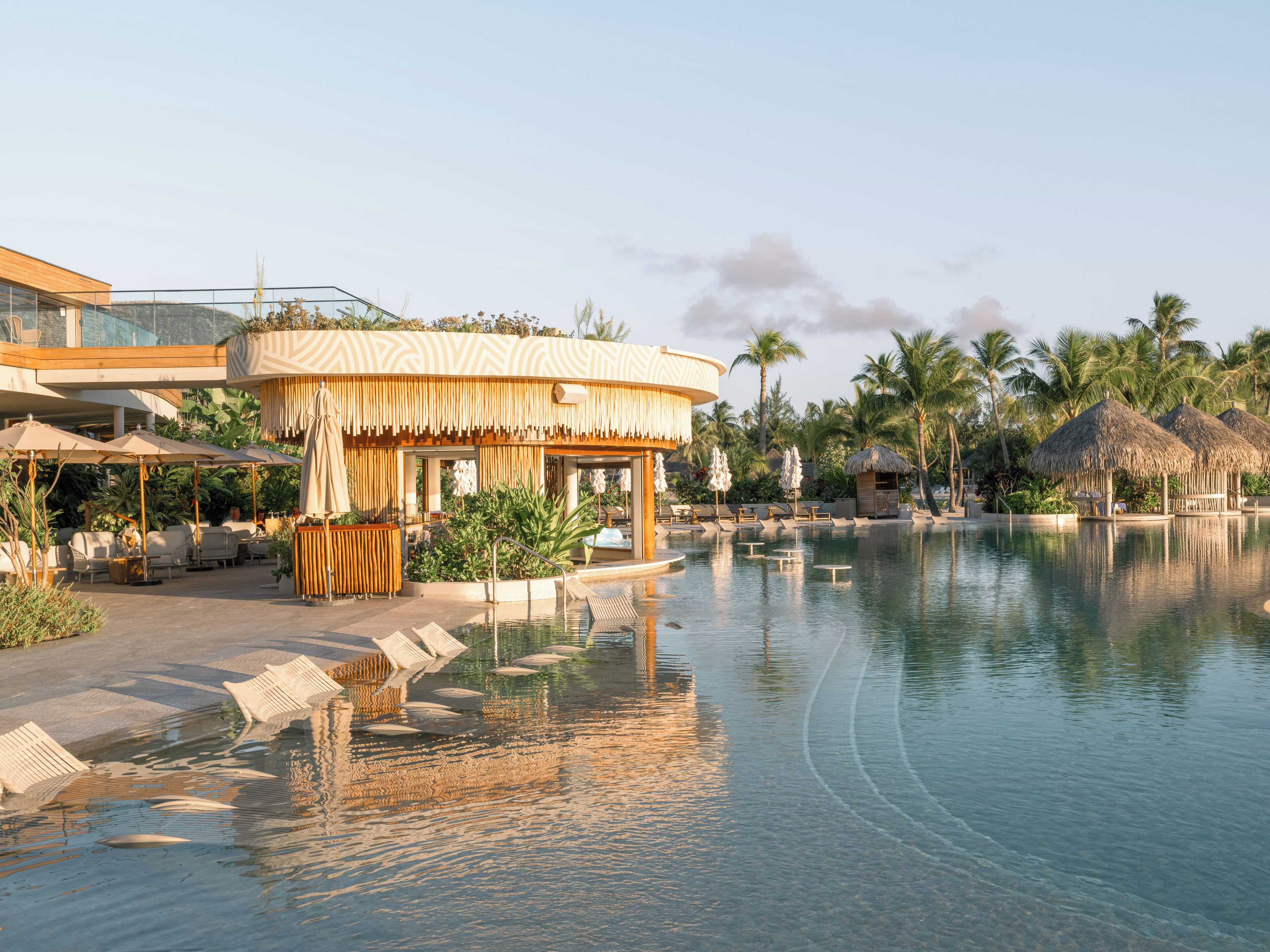 A resort pool area with a thatched-roof structure, lounge chairs, and palm trees.