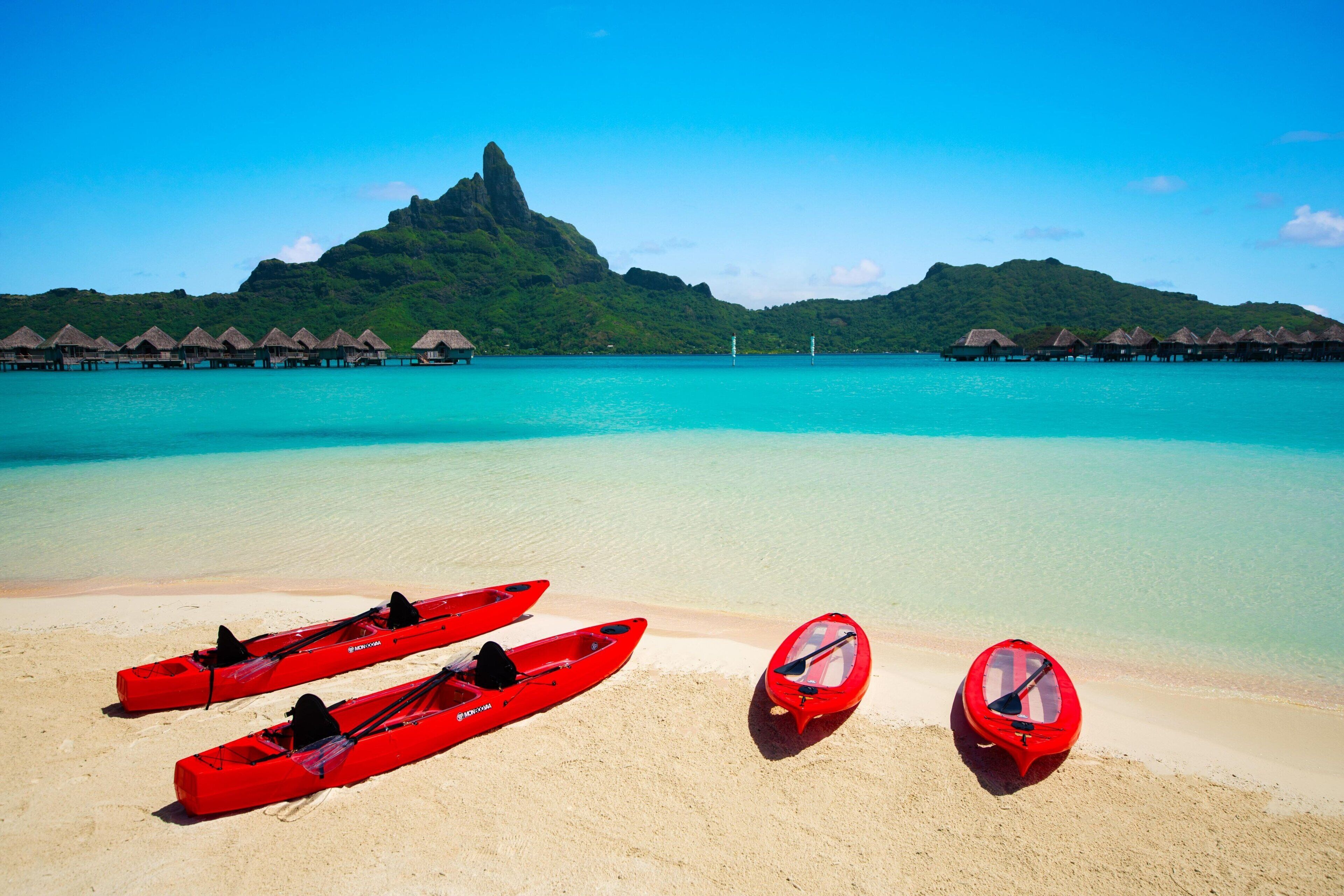A beach with three red kayaks lined up on the sand, a clear turquoise sea, and thatched-roof overwater bungalows in the background.