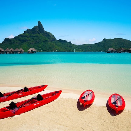 A beach with three red kayaks lined up on the sand, a clear turquoise sea, and thatched-roof overwater bungalows in the background.