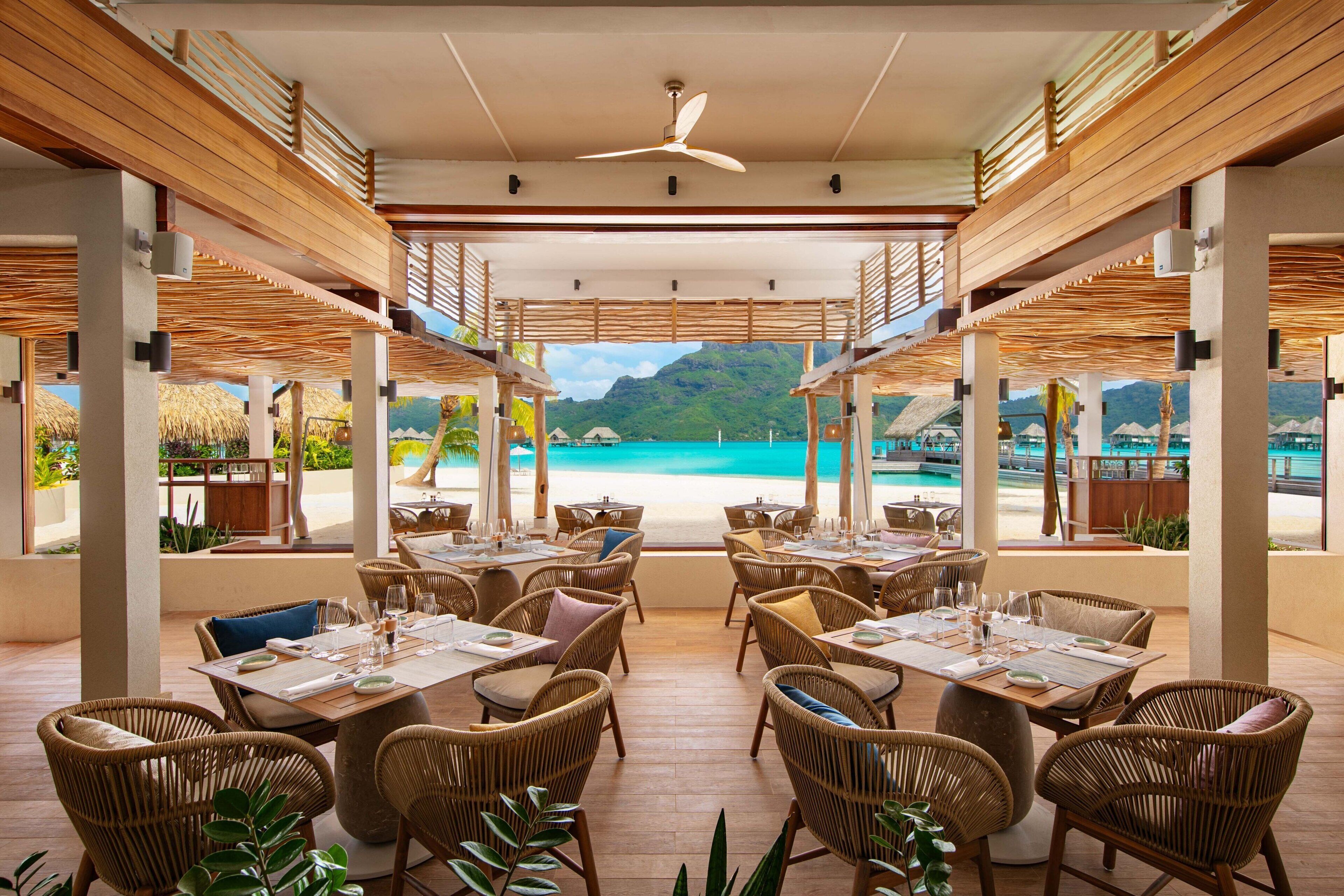 Un restaurante con mesas y sillas de mimbre, con vistas a una playa y montañas.