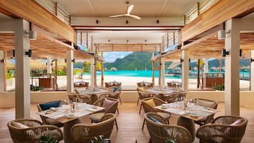 Un restaurante con mesas y sillas de mimbre, con vistas a una playa y montañas.