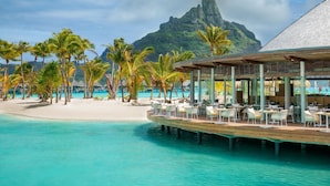 Un complexe hôtelier situé en bord de mer, doté d’une terrasse en bois, de chaises longues et offrant une vue sur une montagne et l’océan.