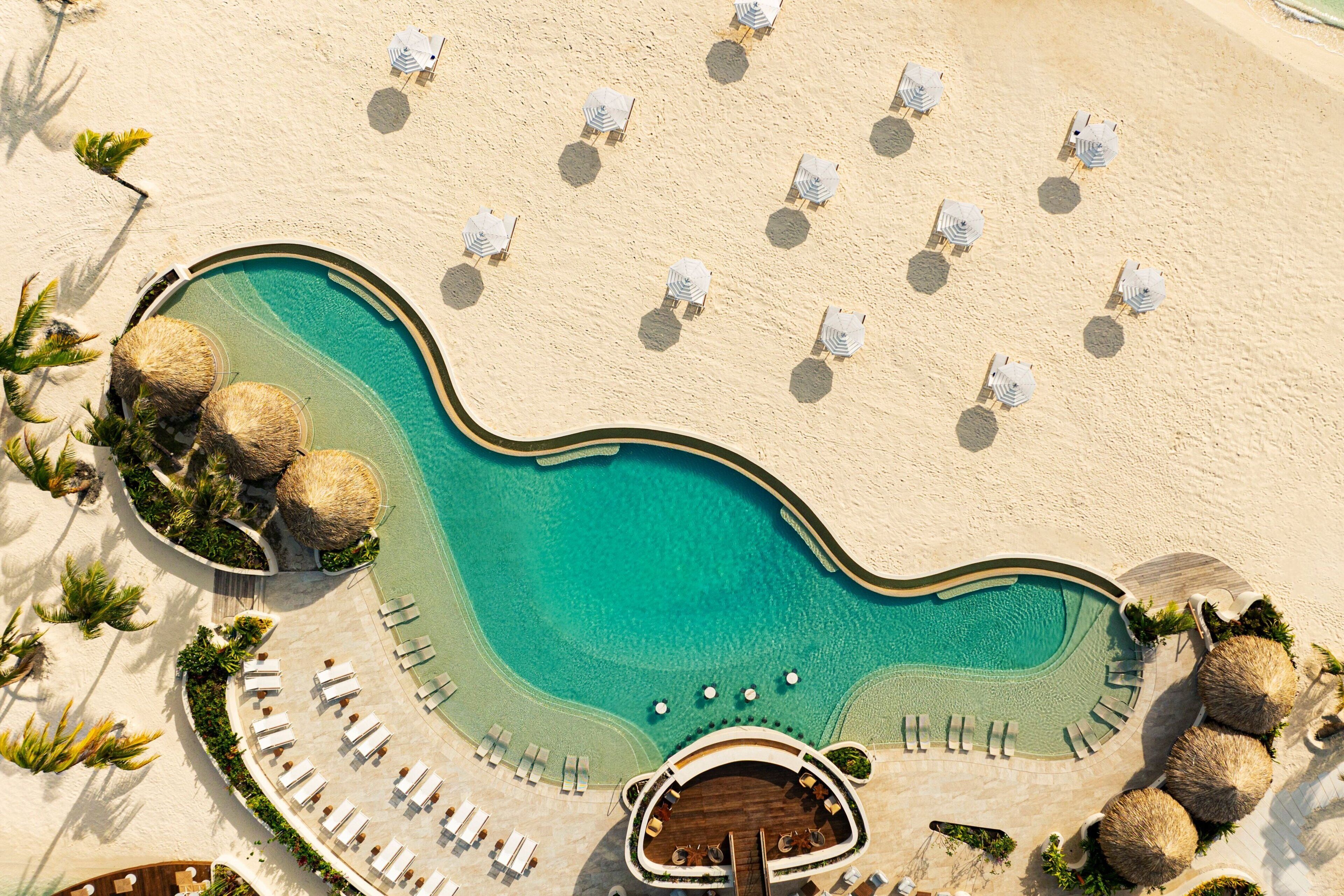 An aerial view of a resort with a large swimming pool, sun loungers, and thatched umbrellas.