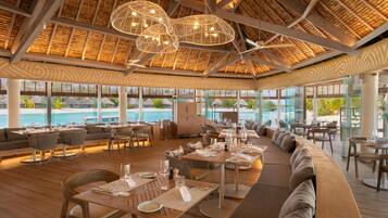 Un amplio espacio exterior para comer con mesas y sillas de madera, techo de paja y grandes ventanas con vista a una piscina.