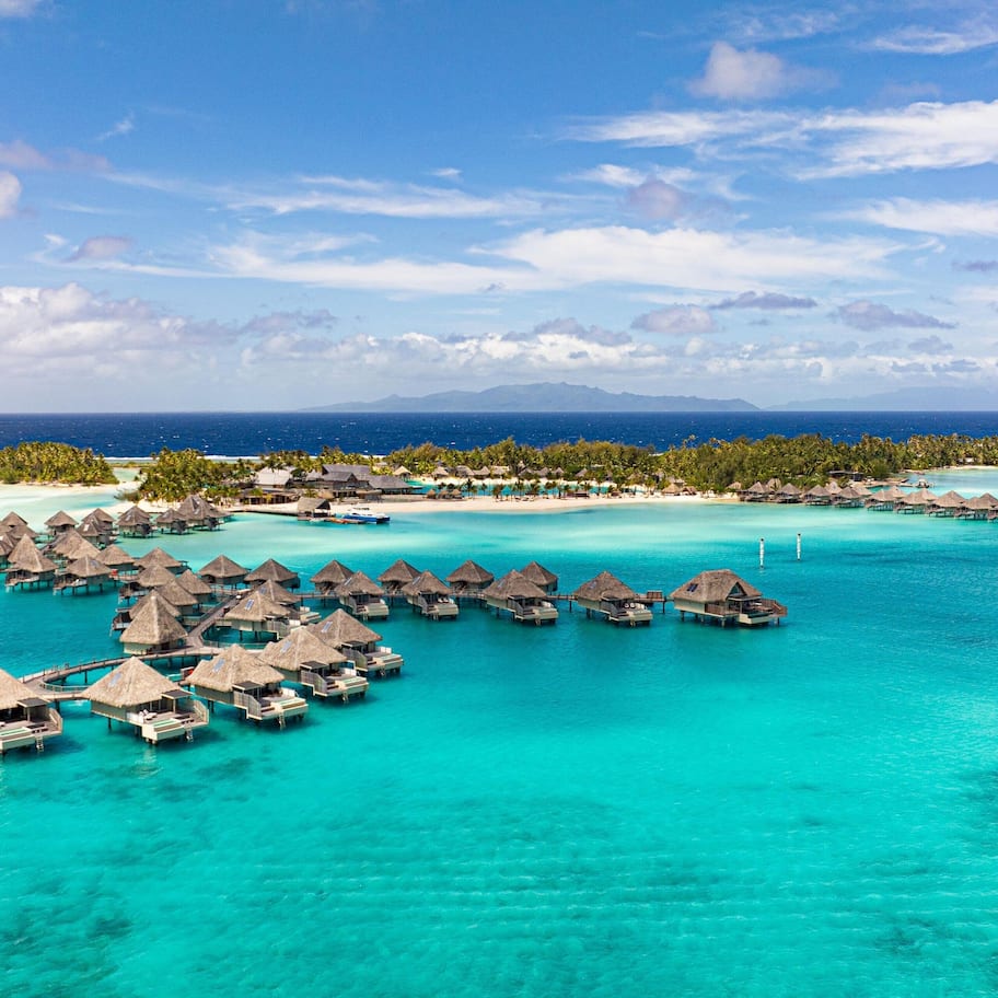 An aerial view of overwater bungalows in turquoise waters, connected by walkways, with a clear sky and distant mountains.