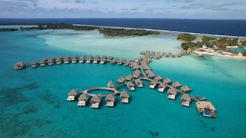 Vista aérea de bungalós sobre el agua dispuestos en forma de semicírculo, unidos por pasarelas, rodeados de aguas turquesas y islas a la distancia.