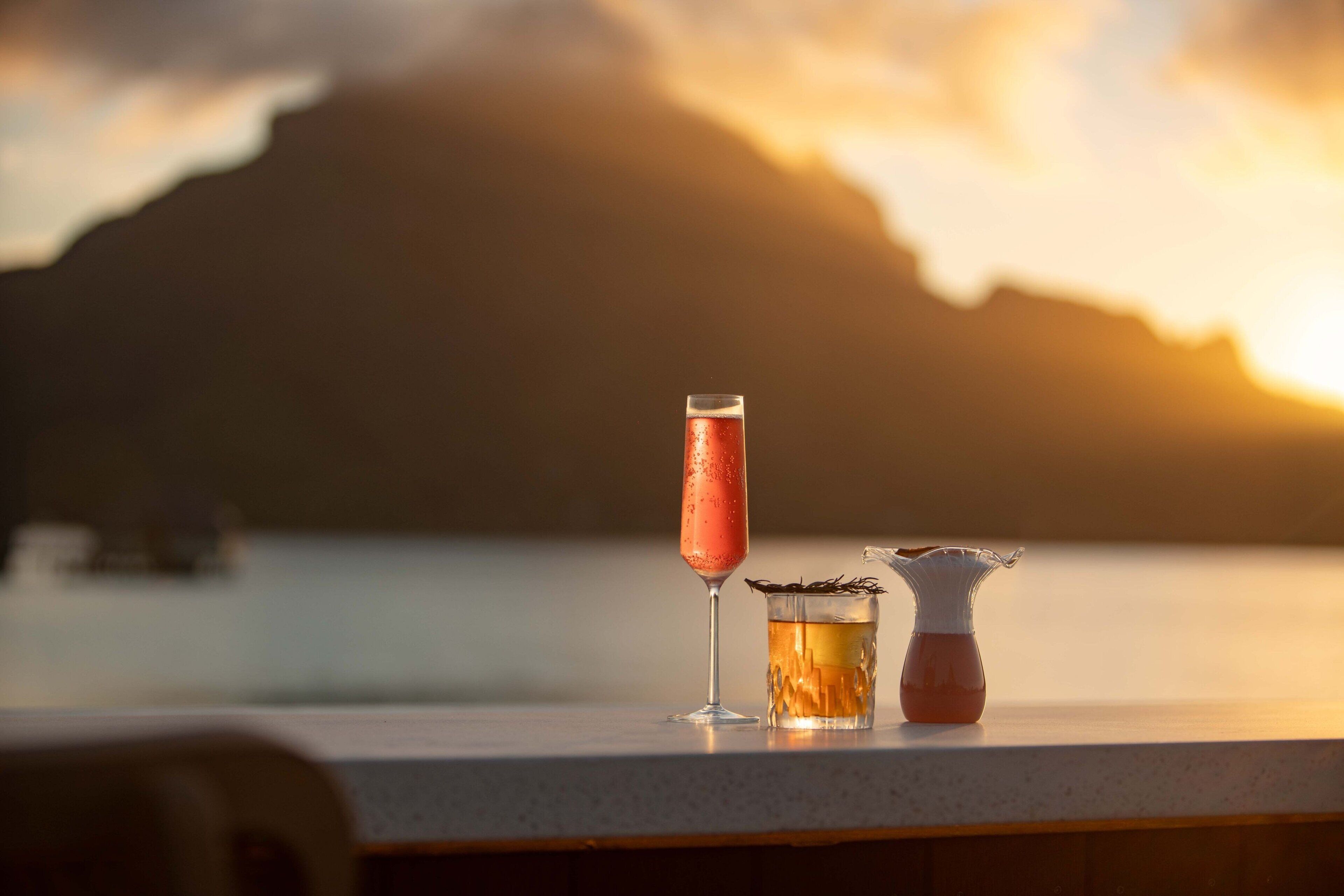 Un bar con un vaso de cóctel y una jarra sobre una barra, con un atardecer de fondo.