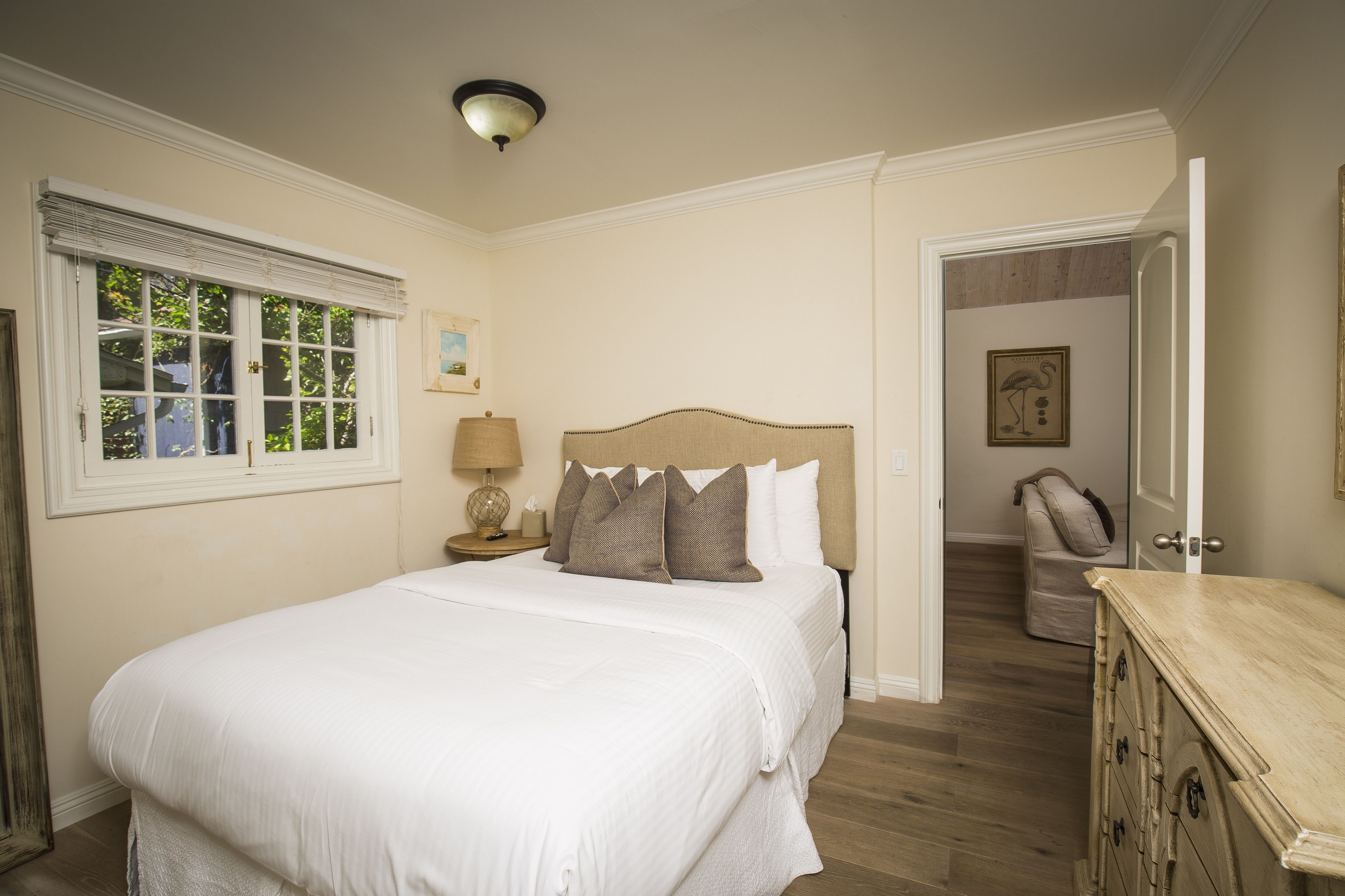 A bedroom with a large bed, a nightstand with a lamp, a door leading to another room, and a dresser.