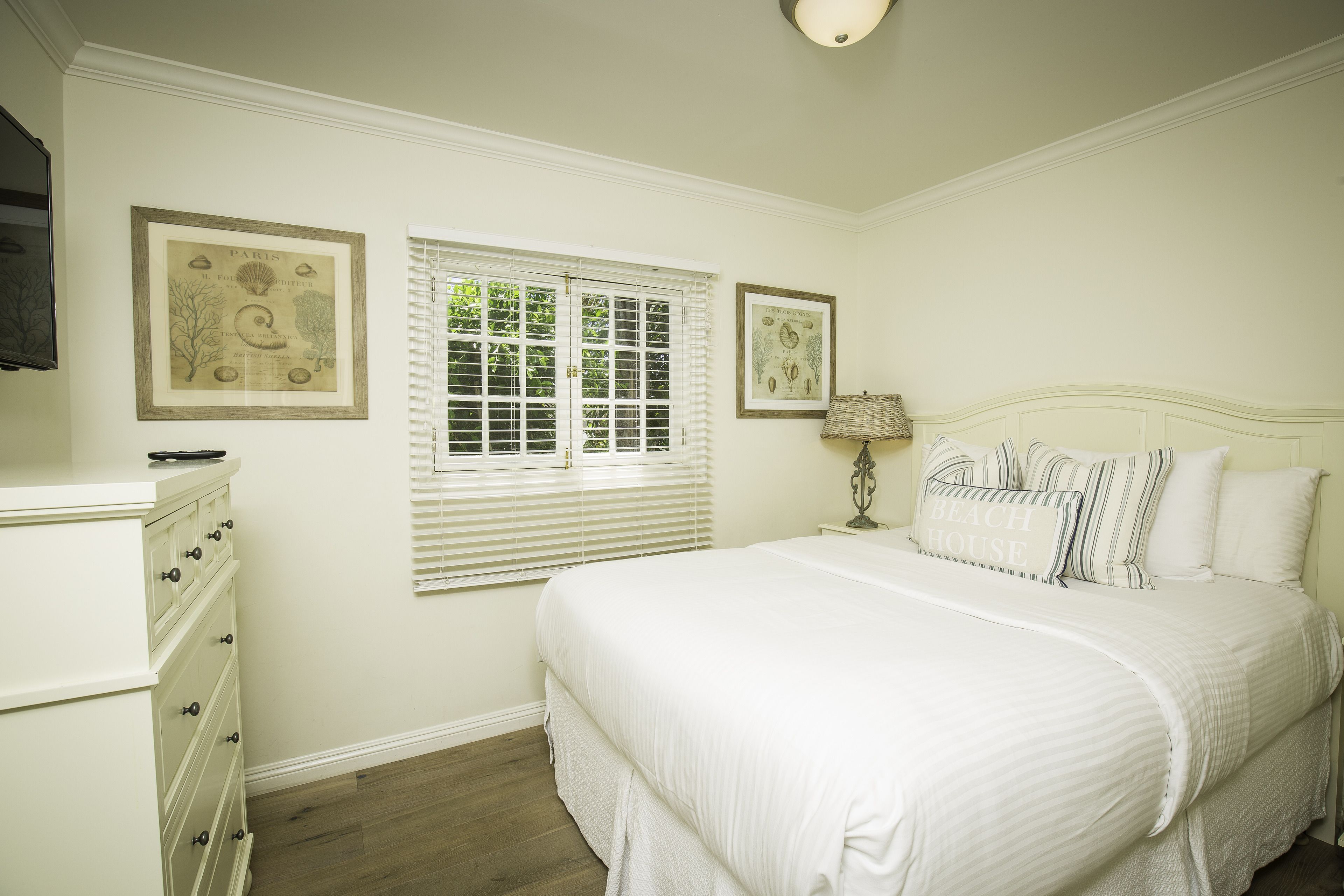 A bedroom with a large bed, a dresser, a window with blinds, and two framed botanical prints on the wall.