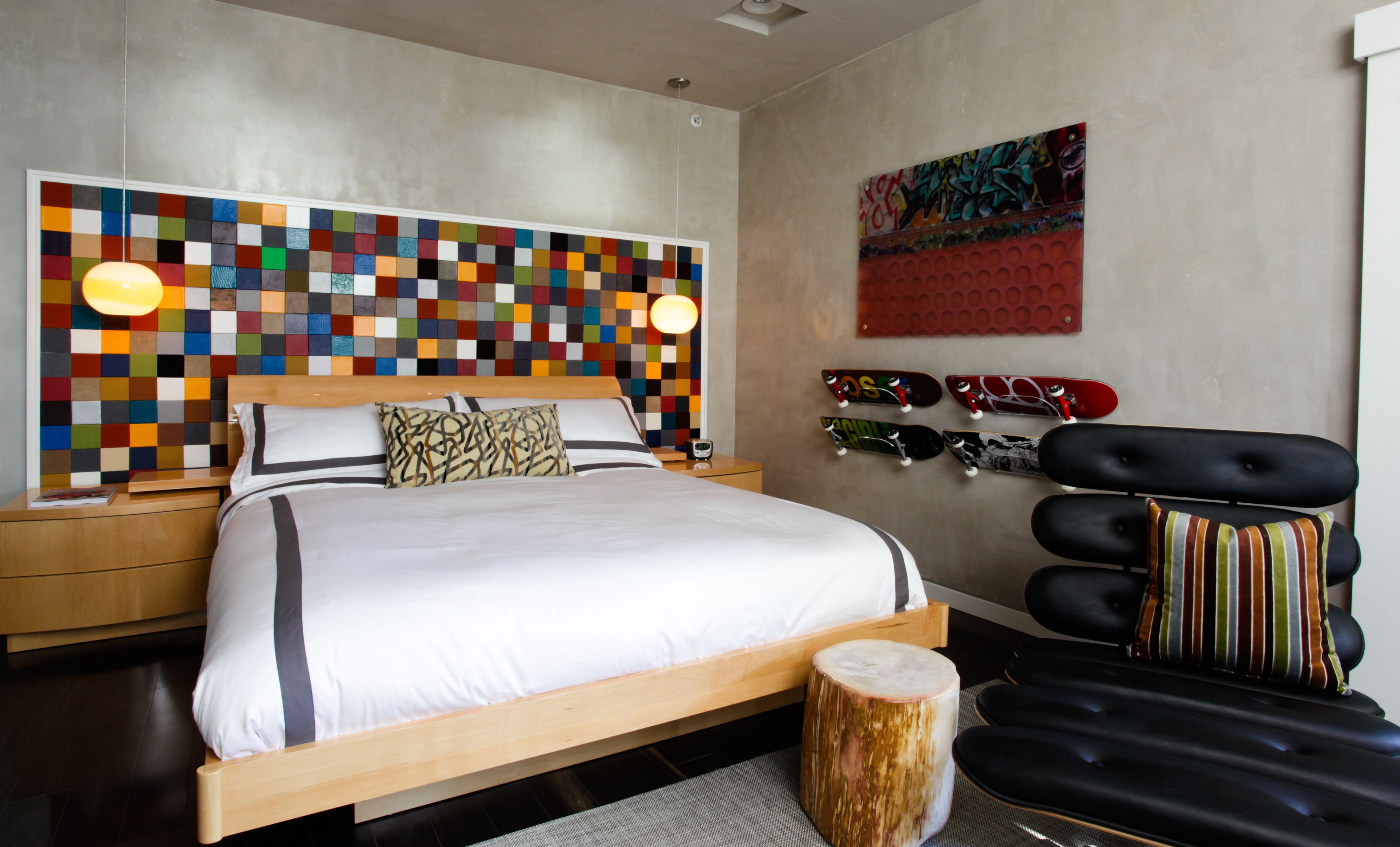 Una camera da letto moderna con una testiera colorata a mosaico, un letto con lenzuola bianche, un comodino in legno, uno sgabello rotondo in legno e una poltrona in pelle nera con cuscini a righe.