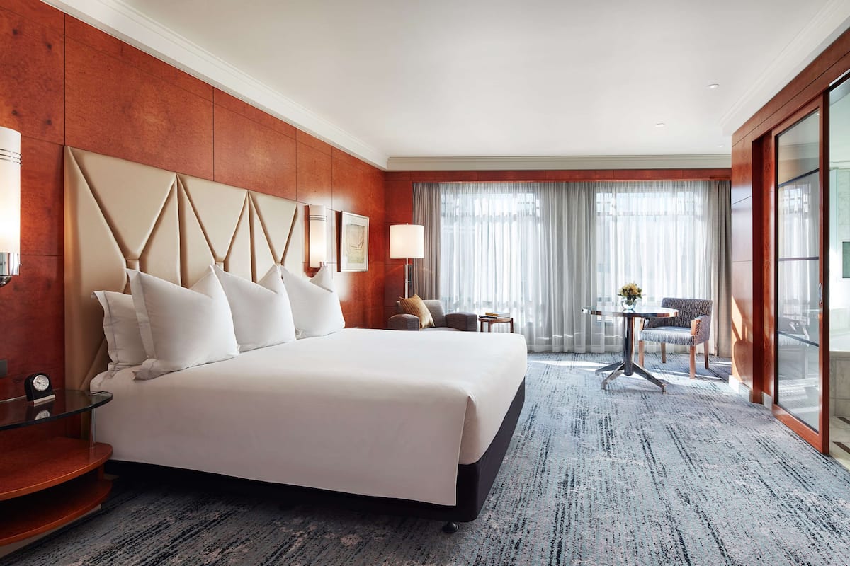A hotel room with a large bed, a small table with a vase, a chair, and a view through a glass door.